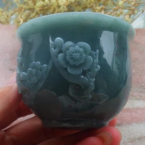 May include: A teal-colored, carved jade cup with floral relief. The cup features a rounded base and a flared rim. The exterior is adorned with detailed carvings of flowers and foliage, showcasing a smooth, polished finish. The cup is held in a hand.