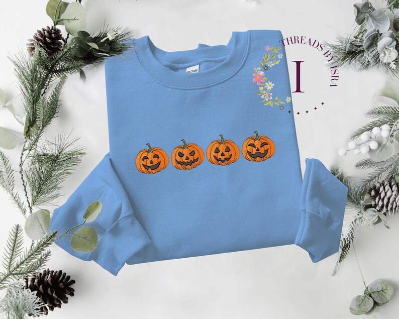 Pu&ograve; includere: Una felpa blu chiaro con una fila di quattro zucche arancioni a forma di jack-o'-lantern con occhi e bocche nere. Le zucche sono ricamate sulla felpa.