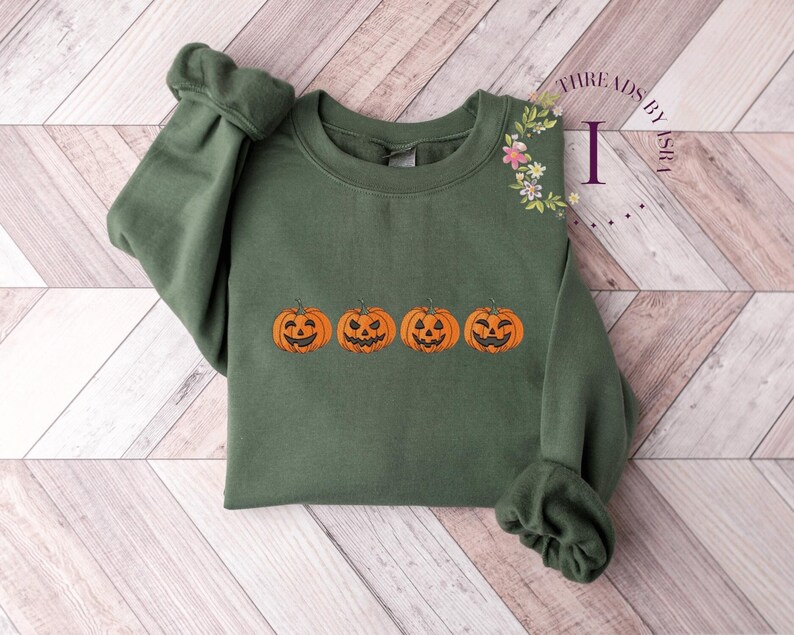 Pu&ograve; includere: Una felpa verde con una fila di quattro zucche arancioni con facce di zucca intagliate ricamate sul davanti. Il testo "THREADS BY ASHLEY" &egrave; ricamato sulla spalla destra.