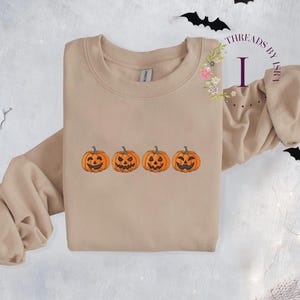 Pu&ograve; includere: Una felpa beige con una fila di quattro zucche arancioni a forma di jack-o'-lantern con diverse espressioni facciali.