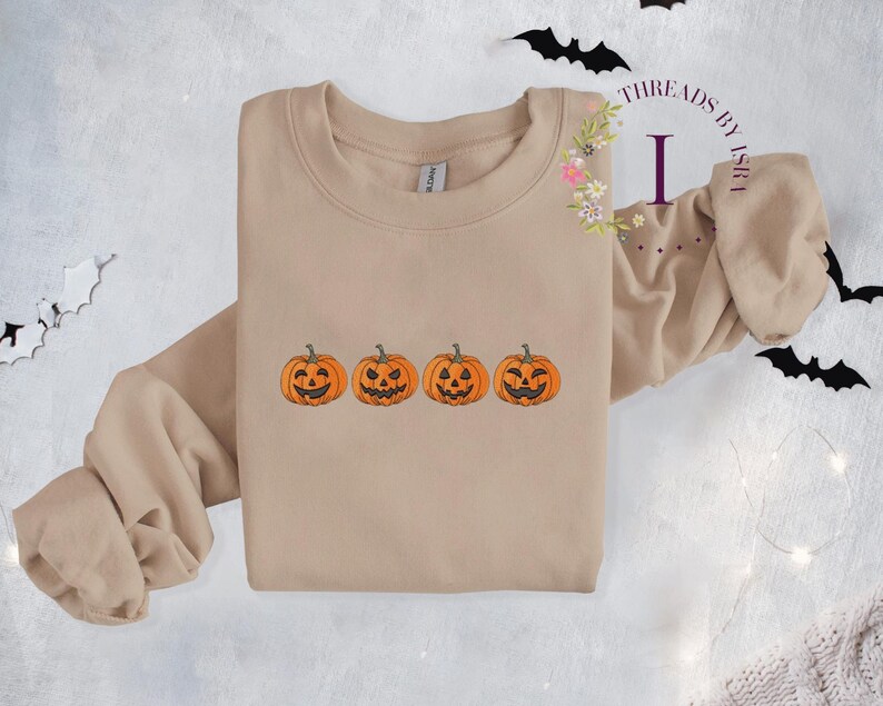 Pu&ograve; includere: Una felpa beige con una fila di quattro zucche arancioni a forma di jack-o'-lantern con diverse espressioni facciali.