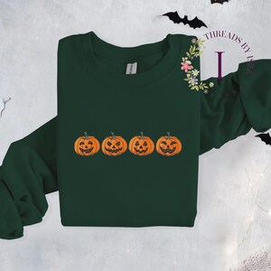 Pu&ograve; includere: Una felpa verde scuro con una fila di quattro zucche arancioni a forma di jack-o'-lantern con facce sorridenti.