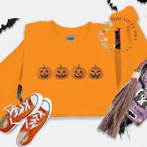 Pu&ograve; includere: Felpa arancione con una fila di quattro facce di zucca intagliate ricamate. La felpa &egrave; su uno sfondo bianco con un paio di scarpe Converse arancioni.