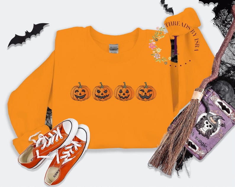 Pu&ograve; includere: Felpa arancione con una fila di quattro facce di zucca intagliate ricamate. La felpa &egrave; su uno sfondo bianco con un paio di scarpe Converse arancioni.