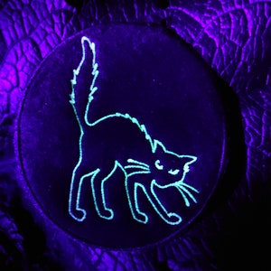 May include: A circular black patch with a glowing green outline of a hissing cat. The cat is arched with its tail up and whiskers visible. The background is a dark purple, textured surface.