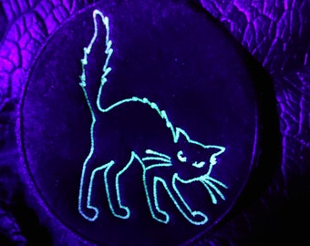 May include: A circular black patch with a glowing green outline of a hissing cat. The cat is arched with its tail up and whiskers visible. The background is a dark purple, textured surface.