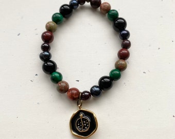 Puede incluir: Un brazalete hecho de cuentas negras, verdes, marrones y rojas con un dije de oro en forma de granada.
