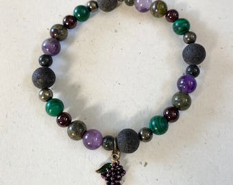 May include: A beaded bracelet with a grape charm. The bracelet is made with black lava rock, green malachite, purple amethyst, and gold pyrite beads.