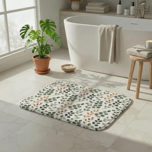 Könnte beinhalten: Eine rechteckige Badematte mit einem botanischen Muster aus grünen und goldenen Blättern auf weißem Grund. Die Matte liegt auf einem gefliesten Boden in einem Badezimmer, mit einer Topfpflanze und einer Badewanne im Blick.