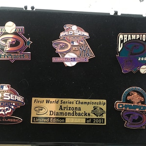 May include: A collection of five Arizona Diamondbacks World Series Championship pins. The pins are displayed in a black display case. The pins feature the team's logo, the year 2001, and the words "World Series Champions". The pins are a limited edition and are numbered 0247 of 2001.