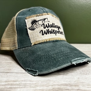 Może przedstawiać: Zielono-beżowa, zniszczona czapka trucker z naszywką z napisem "Walleye Whisperer" i czarno-białym obrazkiem ryby.
