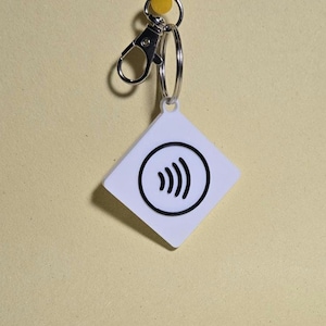 May include: White, diamond-shaped keychain tag featuring a black contactless payment symbol. The tag is attached to a silver metal keyring and clasp. A yellow drawing pin is in the background. The tag is likely used for key identification or payment.