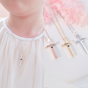 May include: Three gold cross necklaces with personalized engravings. The necklaces feature a small heart or butterfly charm. The engravings include the names "Amelia", "Olivia", and the date "06.21.16".