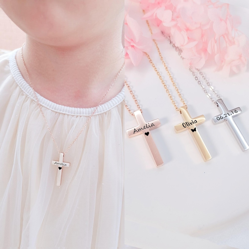 May include: Three gold cross necklaces with personalized engravings. The necklaces feature a small heart or butterfly charm. The engravings include the names "Amelia", "Olivia", and the date "06.21.16".