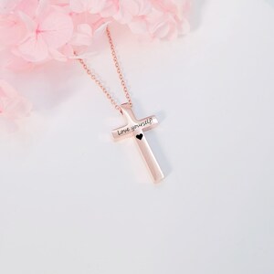 May include: A rose gold cross necklace with the inscription "Love yourself" and a small black heart.