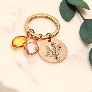 May include: Rose gold keychain with a round charm featuring a floral design and two gemstone charms in yellow and pink. The keychain has a large ring for keys.