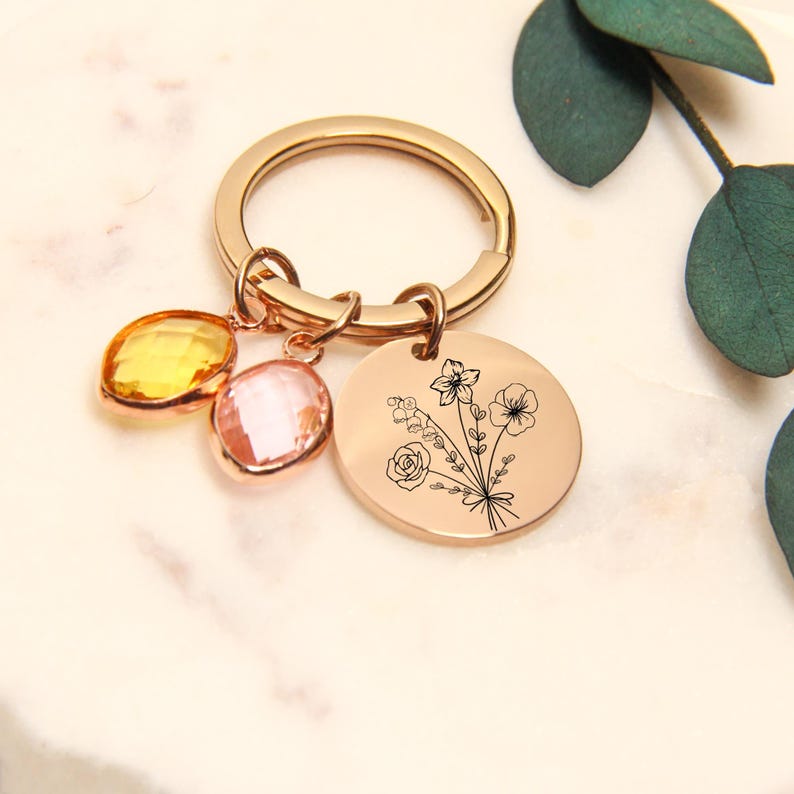 May include: Rose gold keychain with a round charm featuring a floral design and two gemstone charms in yellow and pink. The keychain has a large ring for keys.