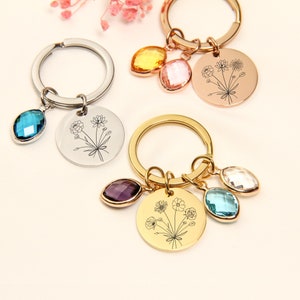 May include: Keychains with floral designs and gemstone accents. One keychain has a silver ring with a blue gemstone and a round pendant. Another has a gold ring with a purple, blue, and clear gemstone and a round pendant.