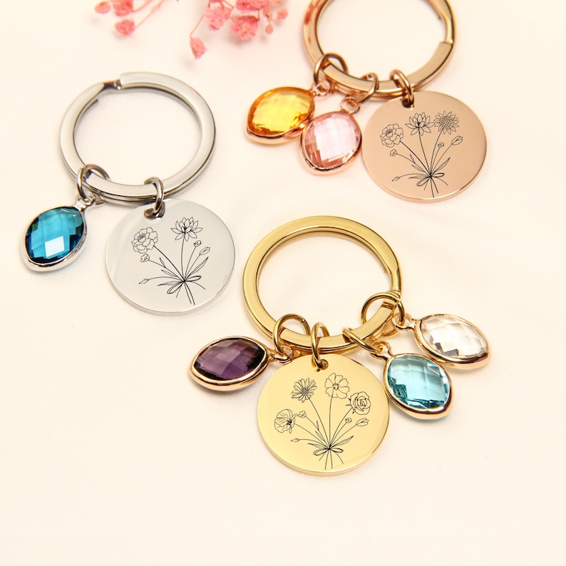 May include: Keychains with floral designs and gemstone accents. One keychain has a silver ring with a blue gemstone and a round pendant. Another has a gold ring with a purple, blue, and clear gemstone and a round pendant.