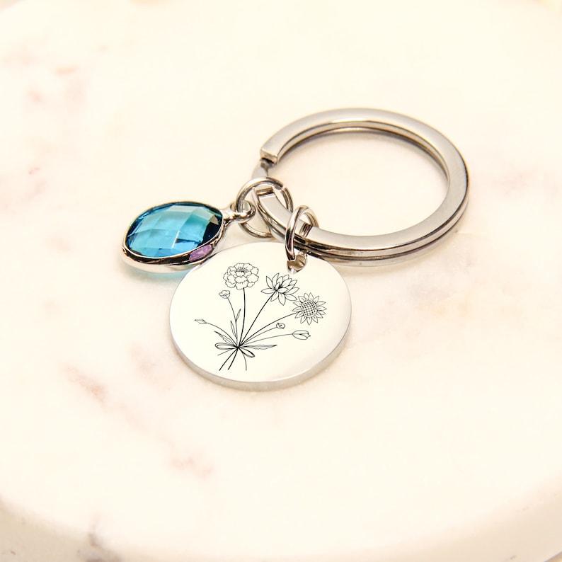 May include: A silver keychain with a round charm featuring a floral design. A blue gemstone hangs from the top. The keychain is on a white marble surface.