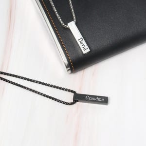 May include: Two silver bar necklaces with black chains. One necklace has the name "David" engraved on the bar and the other has the name "Grandma" engraved on the bar.