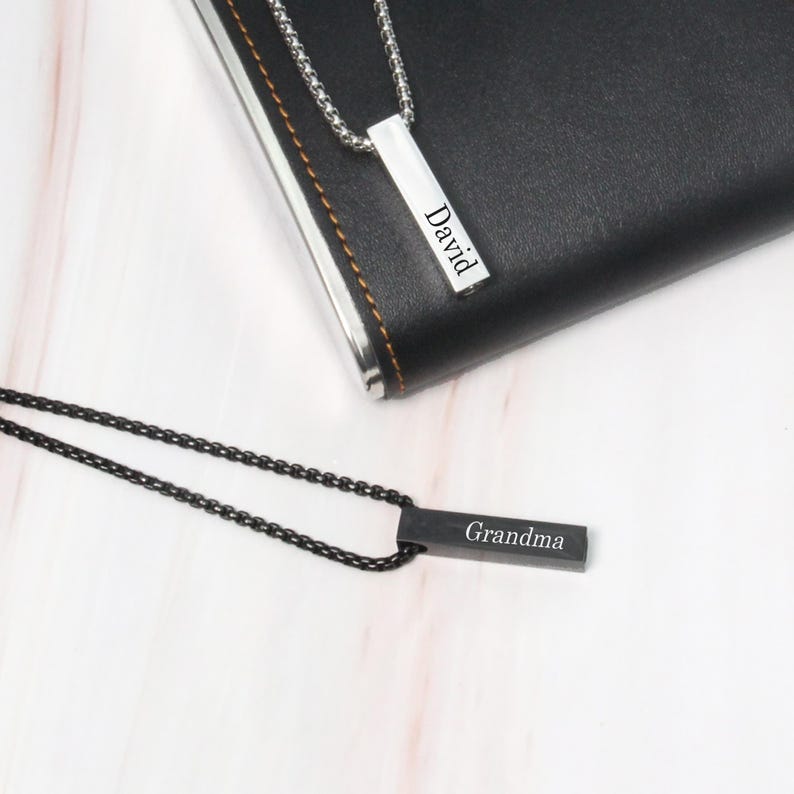 May include: Two silver bar necklaces with black chains. One necklace has the name "David" engraved on the bar and the other has the name "Grandma" engraved on the bar.