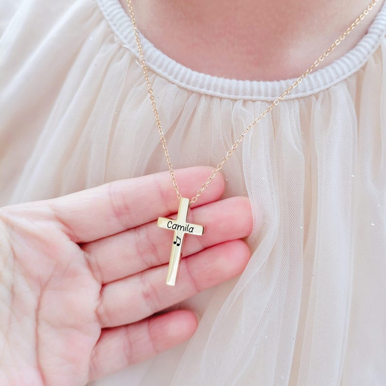 May include: Gold necklace with a cross pendant. The cross has the name "Camila" and a musical note engraved on it.