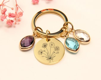 May include: Gold-toned keychain with a round charm featuring a floral design and three gemstone pendants in purple, blue, and clear. The keychain has a large ring for keys and a small ring for the charms.