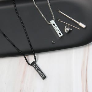 May include: Two silver bar necklaces with black chains. One necklace has the word "DAD" with two hearts on the bar. The other necklace has the Roman numeral date "VIII.II.MMXX" on the bar.
