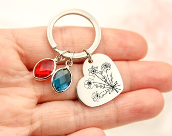 May include: A silver-toned metal keychain featuring a heart-shaped charm engraved with a black line-art floral bouquet. Two marquise-cut faceted crystal charms, one in vibrant red and one in deep blue, are attached to the circular split ring.