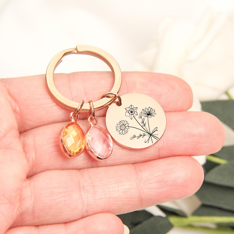 May include: A gold-colored keychain with a round charm featuring a floral design and two teardrop-shaped gemstones in orange and pink. The keychain is held in a hand.