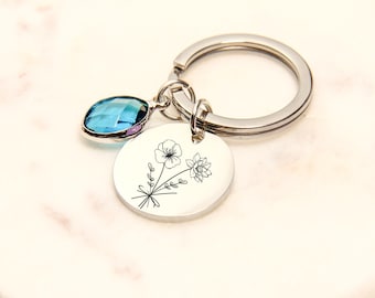 May include: A silver keychain with a round charm featuring a floral design. A blue gemstone and a silver ring are attached to the charm. The keychain is on a white marble surface.