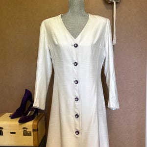 Könnte beinhalten: Ein weißes, langärmliges Kleid mit V-Ausschnitt und lila Knöpfen. Das Kleid wird auf einer Schaufensterpuppe präsentiert, mit einem Paar lila Absätzen und Vintage-Koffern im Vordergrund. Eine Hängepflanze ist im Hintergrund zu sehen.
