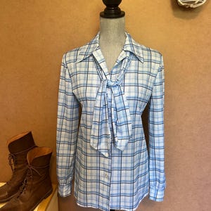 May include: A light blue and white plaid button-up shirt with a necktie. The long-sleeved shirt is displayed on a mannequin. Brown leather boots are visible in the background.