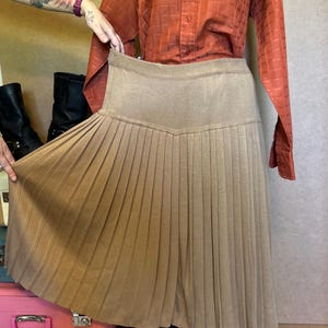 May include: A tan pleated skirt with a ribbed waistband. The skirt is knee-length and has a classic design. The skirt is displayed on a mannequin with a rust-colored button-down shirt.