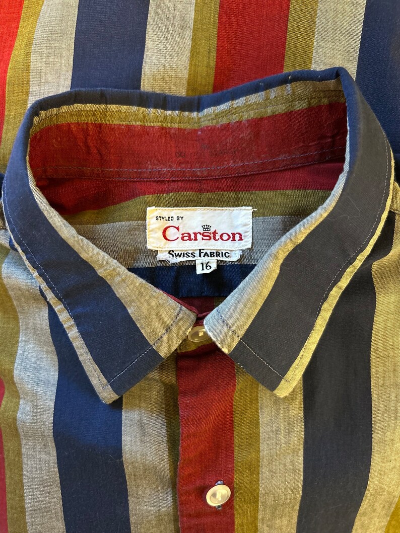 Peut inclure: Une chemise &agrave; boutons ray&eacute;e avec un motif bleu, rouge et vert. La chemise porte une &eacute;tiquette indiquant "Styled by Carston Swiss Fabric 16".