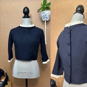 Könnte beinhalten: Marineblaue Vintage-Jacke mit rundem Ausschnitt und langen Ärmeln. Die Jacke hat einen weißen Rüschenkragen und -manschetten sowie ein kleines Blumendetail auf der Vorderseite. Die Jacke wird auf einer Schaufensterpuppe präsentiert.