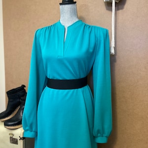 May include: A teal blue long-sleeved dress with a V-neck and a black belt. The dress has a slightly puffed shoulder and a fitted waist.