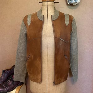 May include: A brown suede bomber jacket featuring grey knit sleeves, a ribbed grey collar, and a front zip fastening. The jacket has two slanted zipped pockets on the front and is displayed on a beige dress form mannequin.