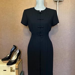 May include: A black dress with short sleeves and a mandarin collar. The dress has a fitted silhouette and features a row of black knotted buttons down the front.