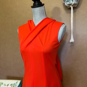 May include: A sleeveless, bright orange dress with a unique draped neckline. The dress is displayed on a mannequin, showcasing its tailored fit and flowing design. The vibrant color and elegant style suggest a vintage or retro aesthetic.