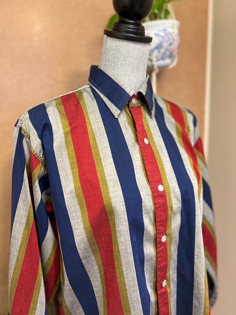 Peut inclure: Une chemise &agrave; boutons ray&eacute;e avec des rayures bleues, rouges, dor&eacute;es et grises. La chemise est sur un mannequin.