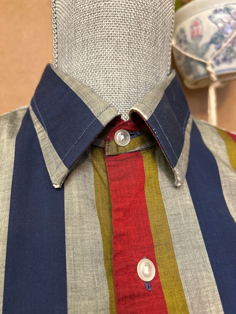 Peut inclure: Une chemise &agrave; boutons ray&eacute;e avec des rayures bleues, rouges, vertes et grises. La chemise a un col et deux boutons.