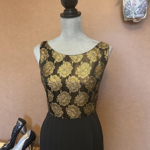 Könnte beinhalten: Schwarzes ärmelloses Kleid mit goldfarbenem floralem Oberteil, präsentiert auf einer Schaufensterpuppe. Schwarze High Heels stehen neben dem Kleid auf einem Vintage-Koffer. Eine dekorative Pflanze hängt im Hintergrund.