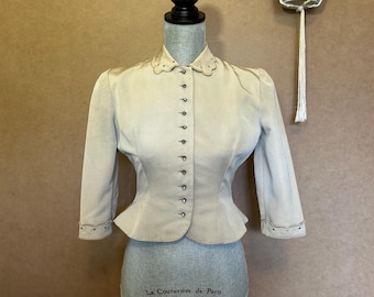 May include: A light beige vintage jacket with a scalloped collar and button-down front. The three-quarter sleeves have decorative stitching. The jacket is displayed on a mannequin with the text "La Couturière de Paris" on the base.