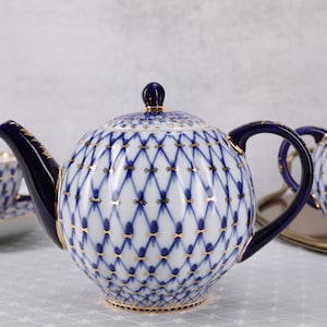 May include: A porcelain tea set featuring a teapot, sugar bowl, and teacup with saucer. The white porcelain is decorated with a blue and gold lattice pattern. The teapot has a dark blue handle and spout.