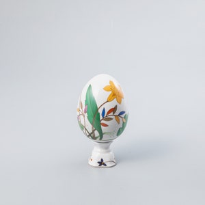 May include: A white ceramic egg with a floral design, featuring a yellow flower and green leaves, stands on a white pedestal. The egg-shaped ornament has colourful accents of blue and red, and is a decorative item.