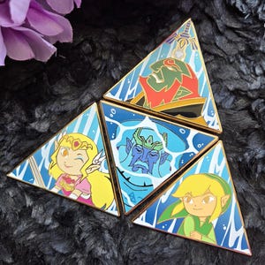 May include: A set of four triangular enamel pins arranged to form a larger triangle. Each pin features a different character illustration with a blue water-like background. The characters include a green-skinned figure, a blonde character, a blue-skinned figure, and a blonde character in green clothing.