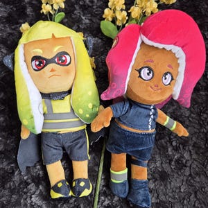 May include: Two plush dolls, one yellow and one pink, resembling characters from a video game. The yellow doll has a black and yellow outfit, while the pink doll wears a blue outfit. Both dolls have detailed facial features and are set against a dark background.