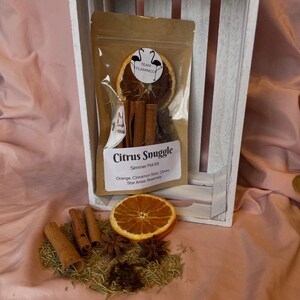 May include: A brown paper bag labeled "Citrus Snuggle" containing a simmer pot kit. The kit includes orange slices, cinnamon sticks, cloves, star anise, and rosemary. The bag is inside a white wooden crate, with some ingredients scattered in front.